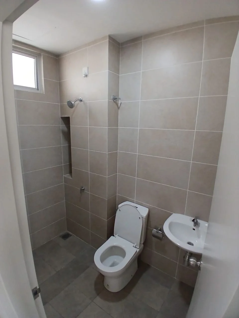 Bathroom features: light beige tile cladding on walls, grey tile flooring, one-piece toilet, wall-mounted basin, chrome basin mixer faucet, shower with wall-mounted shower head, small window for natural light, towel rail, built-in niche for toiletries; style: modern.